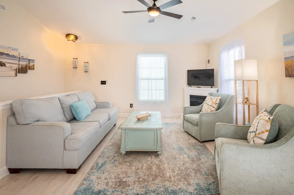A bright, inviting living room featuring a comfortable sofa, a coffee table, several chairs, a large window bringing in natural light, and a television as the centerpiece. The space is accentuated with coastal-themed decor, creating a relaxed and welcoming atmosphere perfect for gatherings or quiet evenings in.