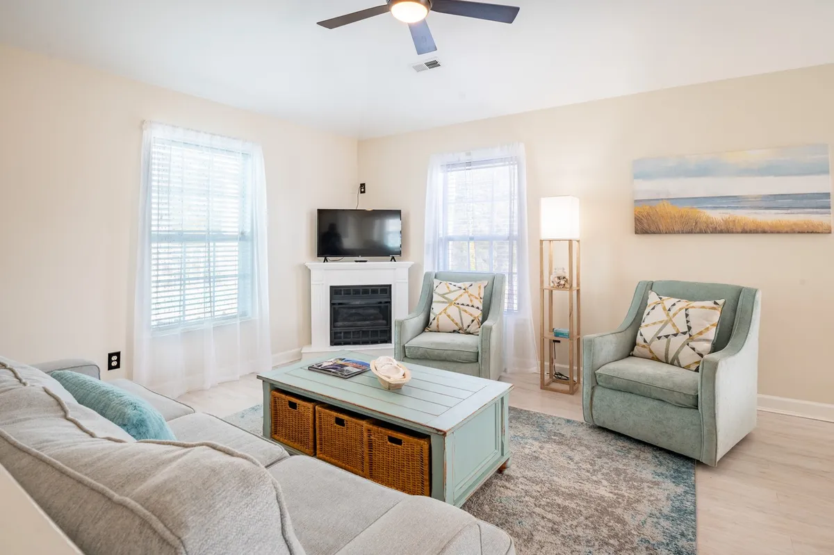 A tranquil, airy living room featuring a sofa, coffee table, armchairs, a television, and coastal artwork. The space feels open and welcoming, encouraging group activities, relaxation, or watching TV together.