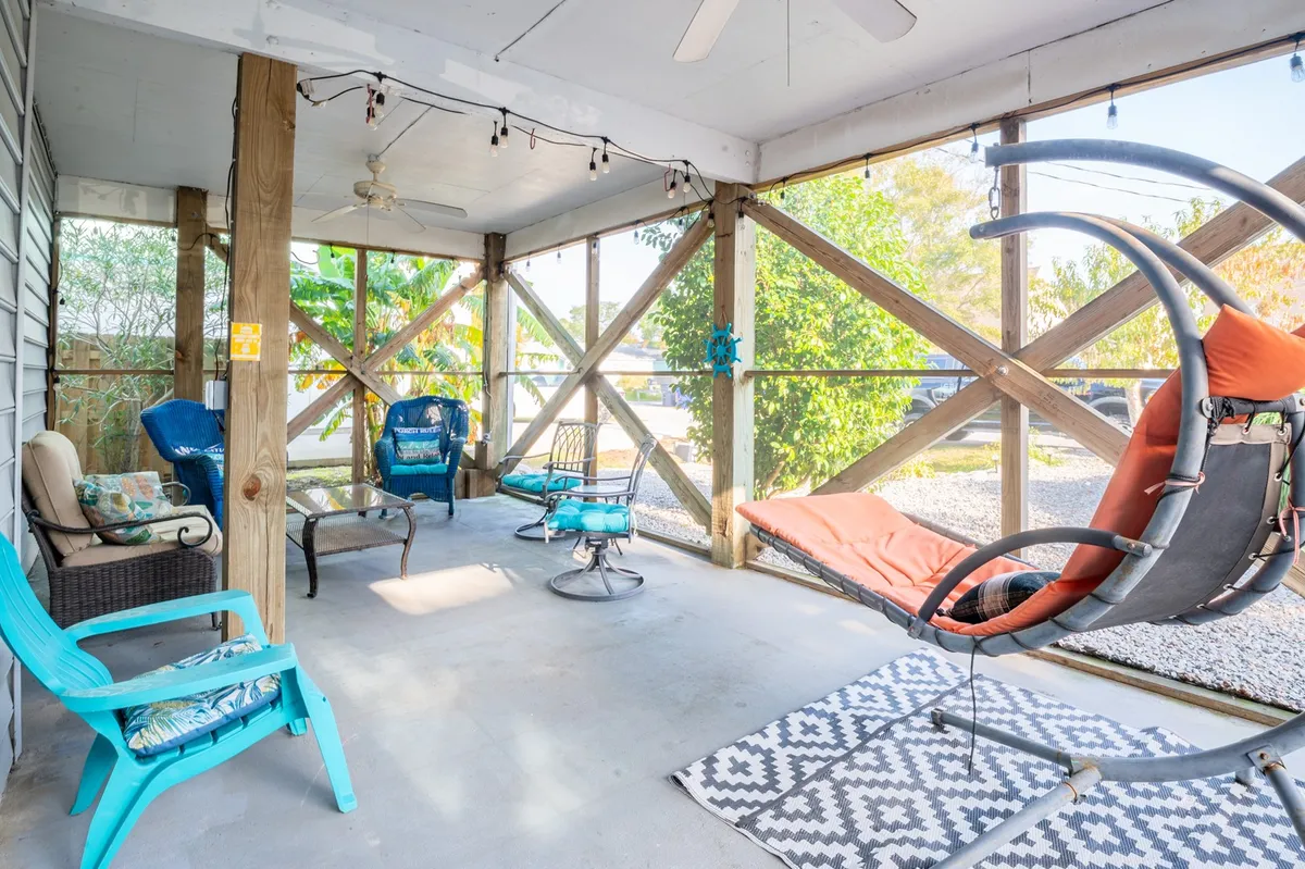 A breezy indoor-outdoor space featuring a hanging chair, colorful and comfortable seating, a decorative rug, and exposed wooden beams. Large windows or doors open to leafy surroundings, blurring the line between inside and outside. The relaxed arrangement suggests a perfect spot for unwinding or sharing stories after a day exploring local attractions.