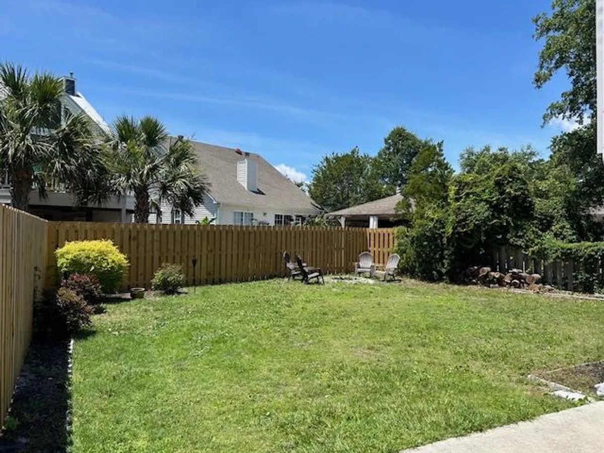 A lush green lawn bordered by a wooden privacy fence, dotted with palm trees and outdoor chairs. The spacious yard offers plenty of room for games or relaxation, perfect for family gatherings or quiet moments in the sun.