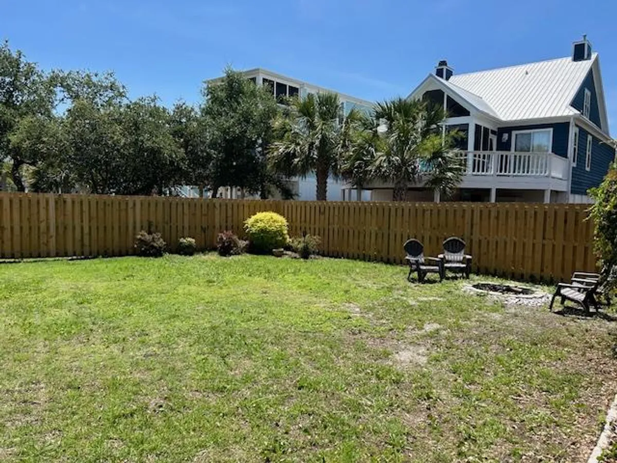 This exterior image showcases a lush green lawn with a seating area featuring chairs, a fenced backyard with palm trees, and the house in the background. The space evokes an inviting, family-friendly atmosphere, perfect for outdoor games, picnics, or casual gatherings after a day at the beach or boardwalk.