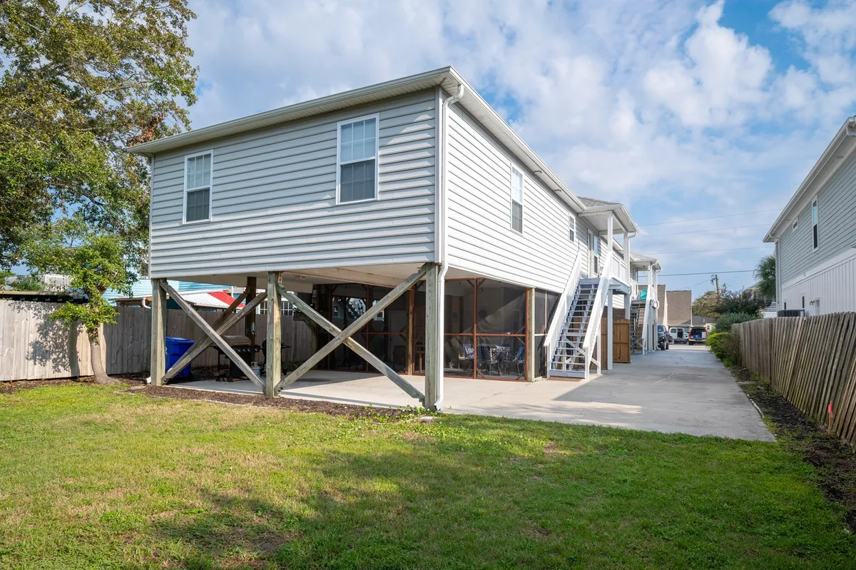 An exterior view of the property features a raised house structure with sturdy pillars, steps leading up, and an open outdoor space that invites gatherings and relaxation. The scene is relaxed and subtly coastal, hinting at beautiful sunset views and the social nature of the area.